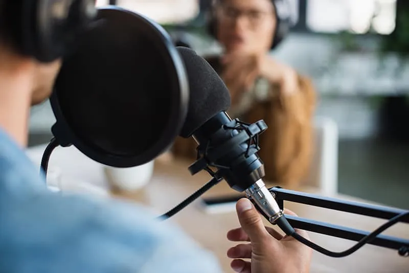 Man wearing headphones in front of a microphone with a blurred person in the background.