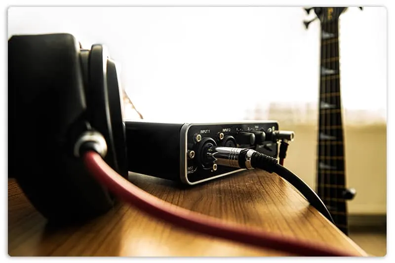 Headphone and soundacard on the table and a bass guitar on the background.