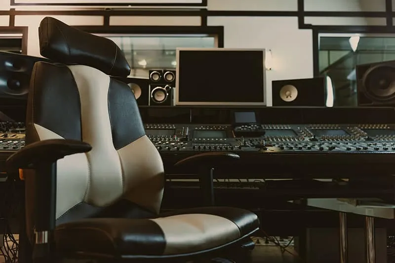 Music studio with a chair in the foreground and a mixing desk in the background with studio monitors.