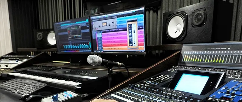 Music studio with screens, studio monitors, mixing desk, keyboard, and microphone.