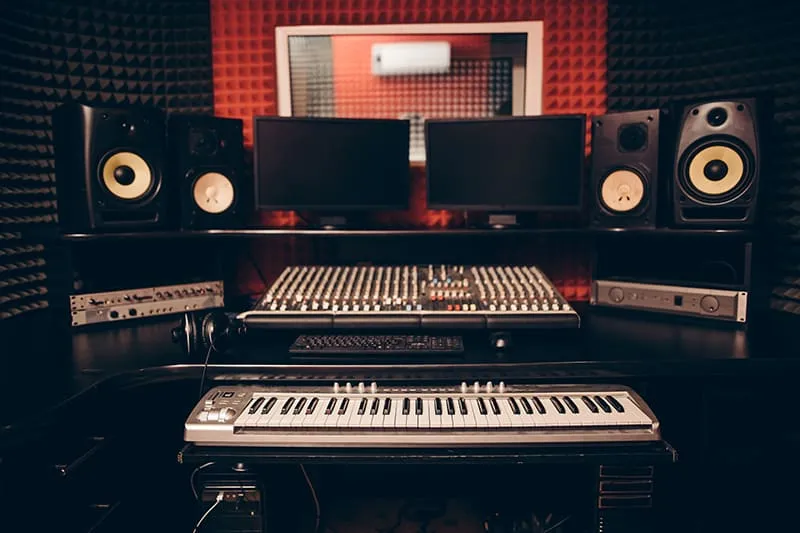 Small recording studio with a keyboard, studio monitors, screen, and mixing console.